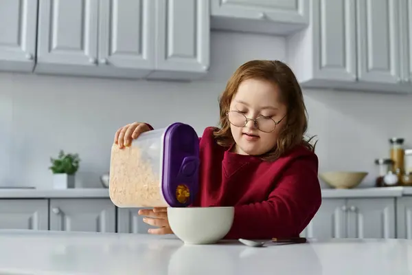Una niña con síndrome de Down vierte cereal en un tazón en su mesa de la cocina. - foto de stock