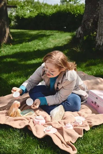 Una niña con síndrome de Down disfruta de una fiesta de té con su muñeca en una manta en el parque. — Stock Photo
