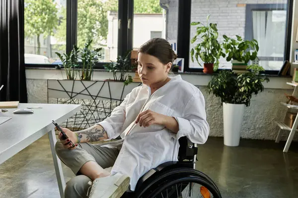 A young businesswoman in a wheelchair uses her phone while sitting at a modern desk in a bright office setting. — Stock Photo