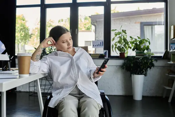 A young businesswoman in a wheelchair works on her phone at a modern office desk, with a coffee cup beside her. — Stock Photo