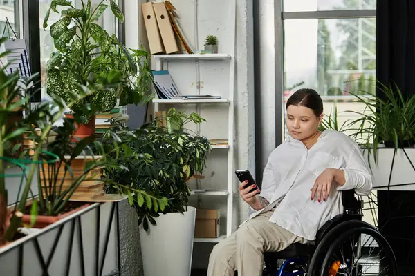 A young businesswoman with a disability sits in a wheelchair and uses her phone in a modern office setting. — Stock Photo