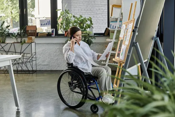 Una giovane donna d'affari con disabilità siede su una sedia a rotelle in un ufficio moderno, lavora a un progetto e parla al telefono.. — Foto stock