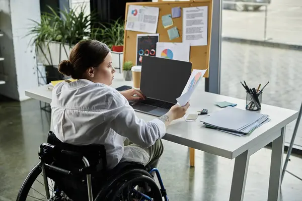 Una joven empresaria en silla de ruedas trabaja en un moderno escritorio de oficina, demostrando su inclusión en el lugar de trabajo. - foto de stock