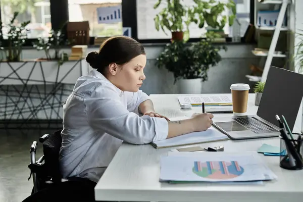 Eine junge Geschäftsfrau im Rollstuhl sitzt an einem Schreibtisch in einem modernen Büro und konzentriert sich auf ihre Arbeit. — Stockfoto