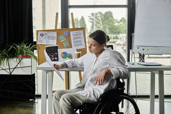 A young businesswoman in a wheelchair sits at a desk in a modern office, reviewing documents. — Stock Photo