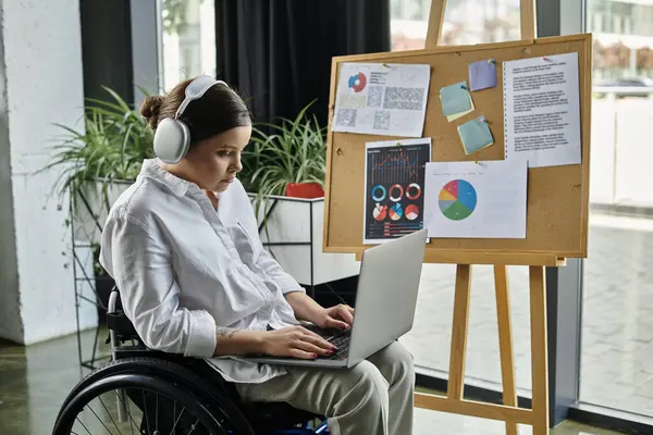 A young businesswoman in a wheelchair works on a laptop in a modern office. She is wearing headphones and appears focused on her task. — Stock Photo