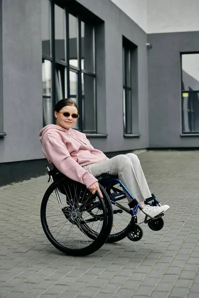 A young woman in a pink hoodie smiles confidently while sitting in her wheelchair outside on a paved area. — Stock Photo
