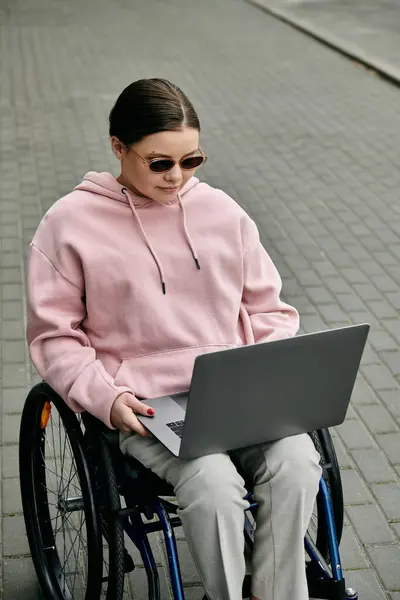 A young woman in a pink hoodie sits in a wheelchair on a paved sidewalk, using a laptop computer. — Stock Photo