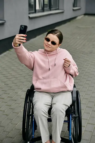 Молодая женщина в розовой толстовке сидит в инвалидном кресле и делает селфи со смартфоном. Она улыбается и выглядит уверенно.. — стоковое фото