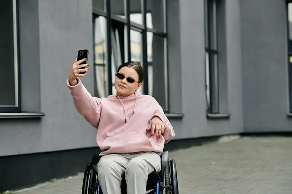 A young woman in a pink hoodie sits in a wheelchair, smiling as she takes a selfie with her smartphone. — Stock Photo
