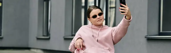 A young woman with disability in a pink hoodie taking a selfie in front of a building. — Stock Photo