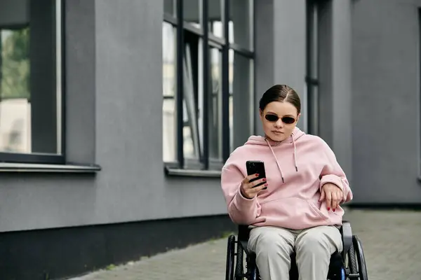 A young woman in a pink hoodie sits in a wheelchair, using her smartphone in a city setting. — Stock Photo