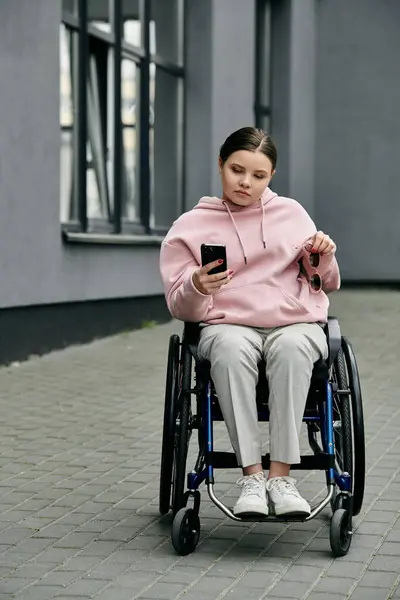 Молодая женщина в розовой толстовке сидит в инвалидном кресле на асфальтированной дорожке, глядя в телефон. — стоковое фото