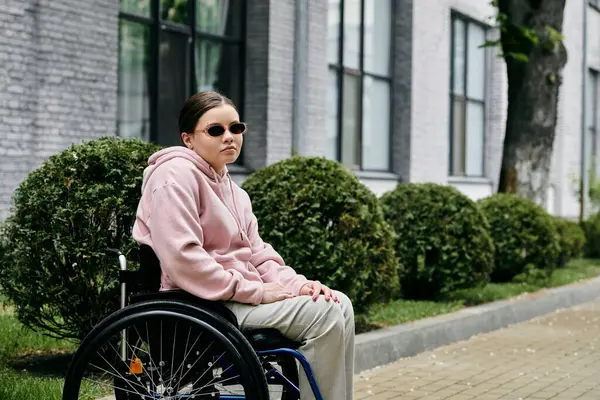 Woman in wheelchair with pink hoodie and sunglasses in front of a building, with bushes and grass in background. — Stock Photo