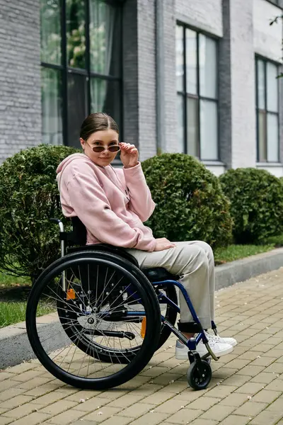 A young woman in a pink hoodie sits in a wheelchair on a sidewalk, adjusting her sunglasses with a confident expression. — Stock Photo