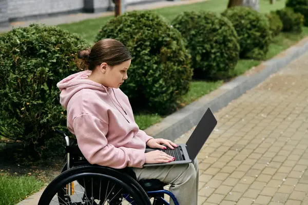 A young woman in a pink hoodie sits in a wheelchair and uses a laptop in a park setting. — Stock Photo