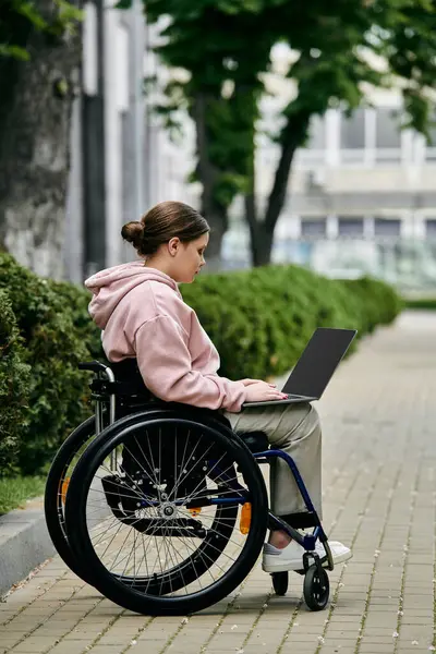 A young woman with a disability sits in a wheelchair on a paved walkway, using a laptop. — Stock Photo