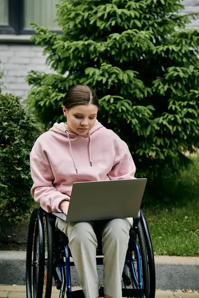 A young woman in a pink hoodie sits in a wheelchair using a laptop outdoors. — Stock Photo