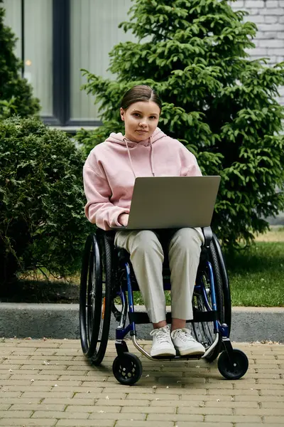A young woman in a pink hoodie sits in a wheelchair outdoors, using a laptop. — Stock Photo