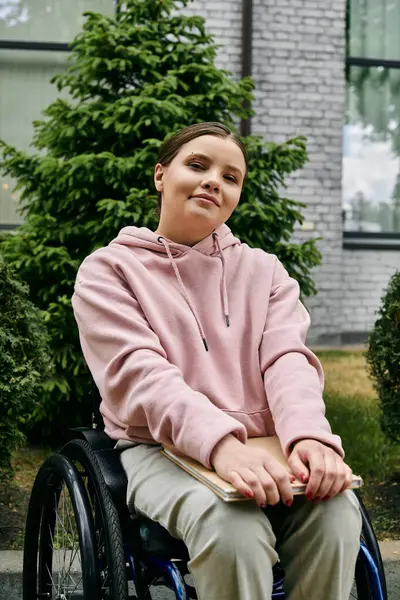 Young woman with disability smiles at camera in pink hoodie, sitting in wheelchair outdoors in a city. — Stock Photo