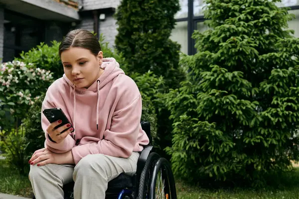A young woman with a disability sits in a wheelchair outside, looking at her phone. — Stock Photo