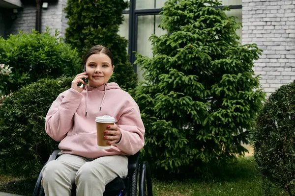 A young woman in a pink hoodie sits in a wheelchair, smiling as she talks on the phone while enjoying a coffee. — Stock Photo