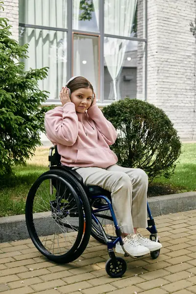 Young woman in pink hoodie relaxes in wheelchair, listening to music on paved walkway. — Stock Photo