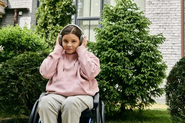A young woman in a pink hoodie sits in a wheelchair outside, listening to music with headphones. — Stock Photo