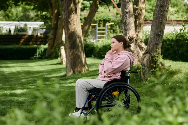 A young woman sits in a wheelchair, thoughtfully gazing at a lush green park. — Stock Photo