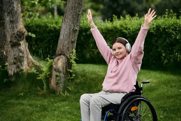A young woman in a pink hoodie sits in a wheelchair and raises her arms in the air as she listens to music with headphones. — Stock Photo