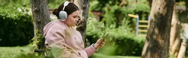 A young woman with a disability sits in a wheelchair outdoors, wearing a pink hoodie and listening to music while using her phone. — Stock Photo