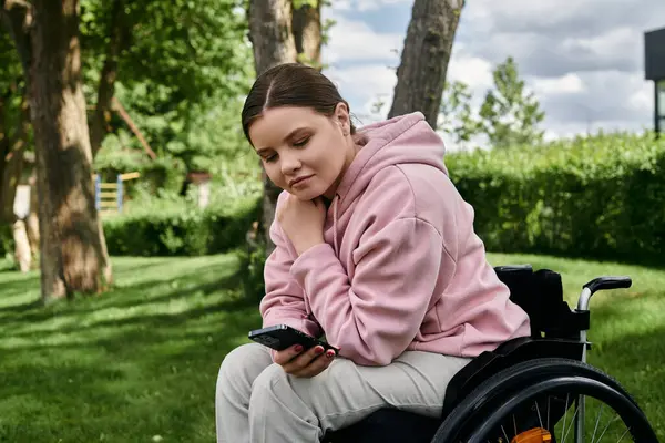 A young woman in a wheelchair sits in a park, looking at her phone. — Stock Photo