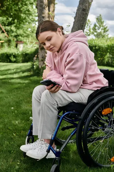 A young woman sits in a wheelchair in a grassy park, looking down at her phone. — Stock Photo