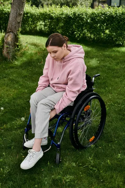 A young woman sits in a wheelchair in a grassy park wearing a pink hoodie. — Stock Photo