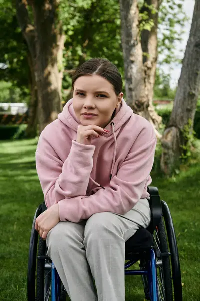 A young woman with a disability sits in a wheelchair, wearing a pink hoodie. She is seated in a park setting. — Stock Photo