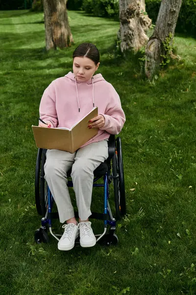 Une jeune femme avec un sweat à capuche rose est assise dans un fauteuil roulant sur une pelouse herbeuse, écrivant dans un carnet. — Photo de stock