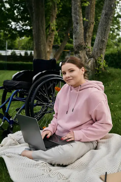 A young woman sits in a wheelchair, using a laptop on a blanket in a park. — Stock Photo