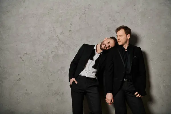 Two men in elegant black suits stand against a grey wall, showing their affection for each other. — Stock Photo