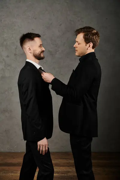A dapper couple in black suits, one adjusting the others tie, standing before a gray wall. — Stock Photo