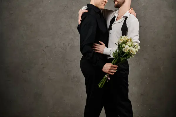 A dapper gay couple embraces, one holding a bouquet of flowers, against a textured concrete wall. — Stock Photo