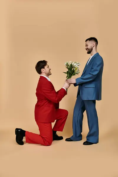 Un couple gay en costumes élégants, un sur un genou avec des fleurs, sur un fond neutre. — Photo de stock