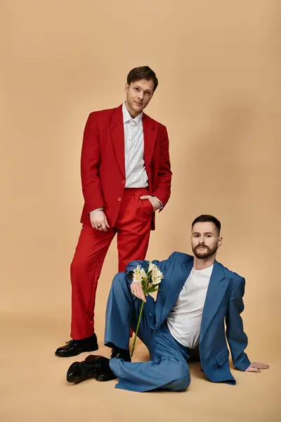 Two men in elegant suits pose: one in red standing tall, the other in blue sitting with flowers. — Stock Photo