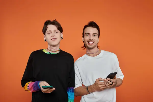 Two young queer friends dressed in stylish, vibrant clothes smile for the camera in front of an orange background. — Stock Photo