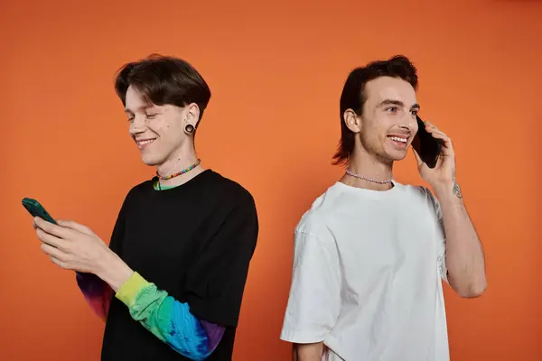 Two stylishly dressed queer friends against a vibrant orange background; one texting while the other is on a call. — Stock Photo