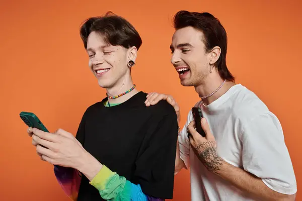 Two young friends, dressed in vibrant attire, pose against an orange backdrop, sharing a laugh while looking at a smartphone. — Stock Photo