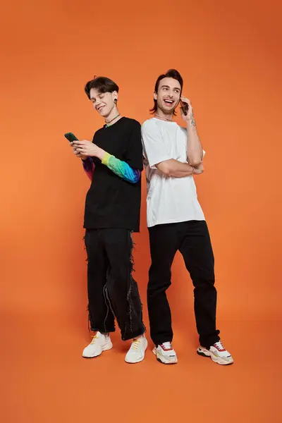 Two stylishly dressed queer friends stand against an orange backdrop, one using a phone while the other chats on one. — Stock Photo