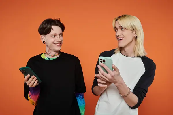 Two young friends in stylish attire stand against a bright orange background, smiling and looking at their phones. — Stock Photo