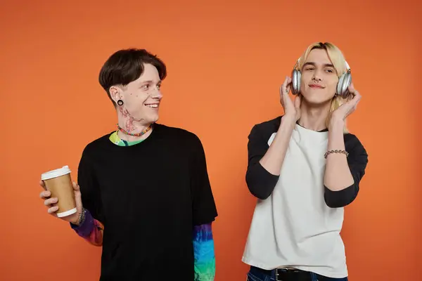 Two young friends, dressed in stylish attire, stand against an orange background, one holding coffee and the other listening to music. — Stock Photo