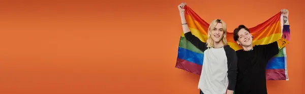Dos jóvenes amigos se paran juntos frente a un fondo naranja, sonriendo y sosteniendo una bandera de orgullo. — Stock Photo