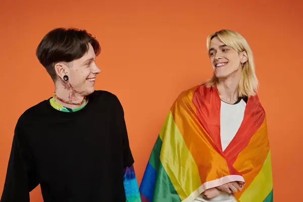 Dos jóvenes queer con la bandera del arco iris orgullo sobre un fondo naranja. — Stock Photo